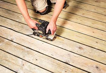 The image shows a person sanding a wooden deck in a backyard patio.